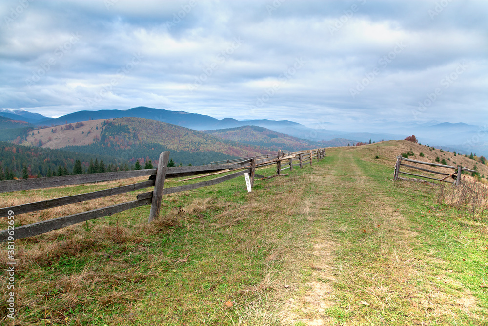 Autumn in the mountains, road with a wooden fence in the foreground, bright mountain slopes on the horizon.