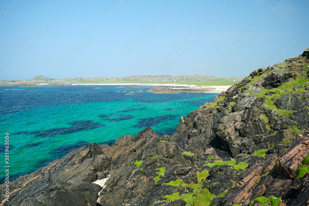 A vibrant view over a rocky coastline along the Isle of Iona, Scotland. Colourful green grasses and a bright blue Atlantic Ocean dominate the image. A sandy beach bay is visible in the background.