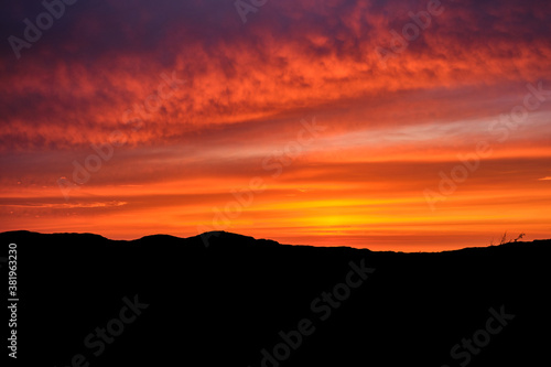 A fiery sky is lit up by the late sunset tones on a summers evening on the Isle of Mull, Scotland. Silhouetted mountains are visible at the bottom of the image and orange and reds light up the sky.