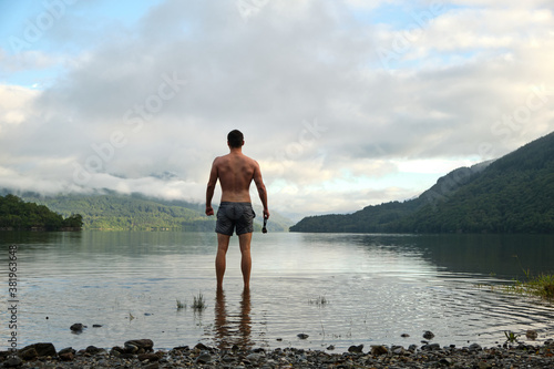 A muscular mid 20s male stands confidently after wild swimming in Loch Lomond, Scotland. Wearing swimming trunks and with swimming goggles in hand. Picturesque views all around. Relaxing wide shot.