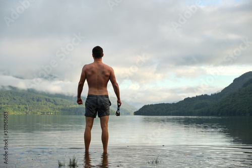 A muscular mid 20s male stands confidently after wild swimming in Loch Lomond, Scotland. Wearing swimming trunks and with swimming goggles in hand. Picturesque views all around. Relaxing.