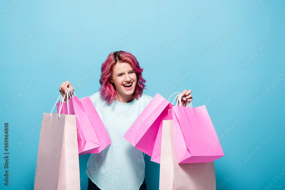 excited young woman with pink hair holding shopping bags on blue background