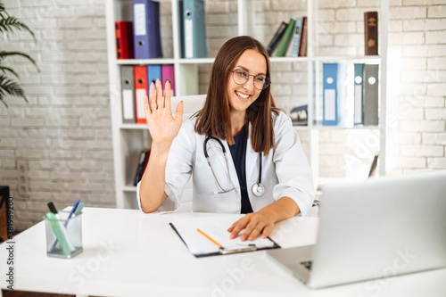 Confident female doctor in eyeglasses sits in the office and using laptop for online consultation. A young woman in white formal coat looks at webcam and waving. Virtual visit concept
