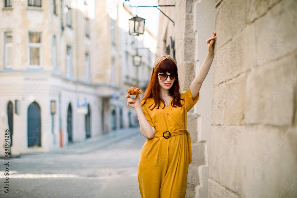 Fashion city portrait of gorgeous young Caucasian red haired woman, dressed in stylish yellow overalls, sunglasses and head hoop, walking in old European city with fresh tasty croissant
