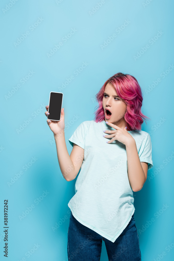 shocked young woman with pink hair holding smartphone with blank screen on blue background