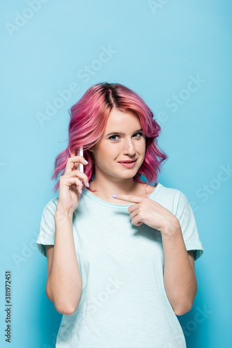 young woman with pink hair pointing at smartphone on blue background