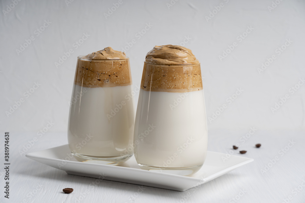 Homemade dalgona coffee made by whipping instant coffee powder, sugar and hot water with addition of cold milk served in drinking glass on rectangular plate on white wooden table. Horizontal Homemade dalgona coffee made by whipping instant coffee powder, sugar and hot water with addition of cold milk served in drinking glass on rectangular plate on white wooden table. Horizontal