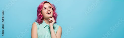 dreamy young woman with pink hair smiling and looking away isolated on blue, panoramic shot