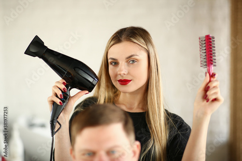 Blonde Hairstylist Showing Blow Dryer and Comb. Woman Hairstylist Using Hairbrush and Hairdryer for Styling Male Haircut in Beauty Salon. Female Stylist Making Hairdo for Client Looking at Camera Shot