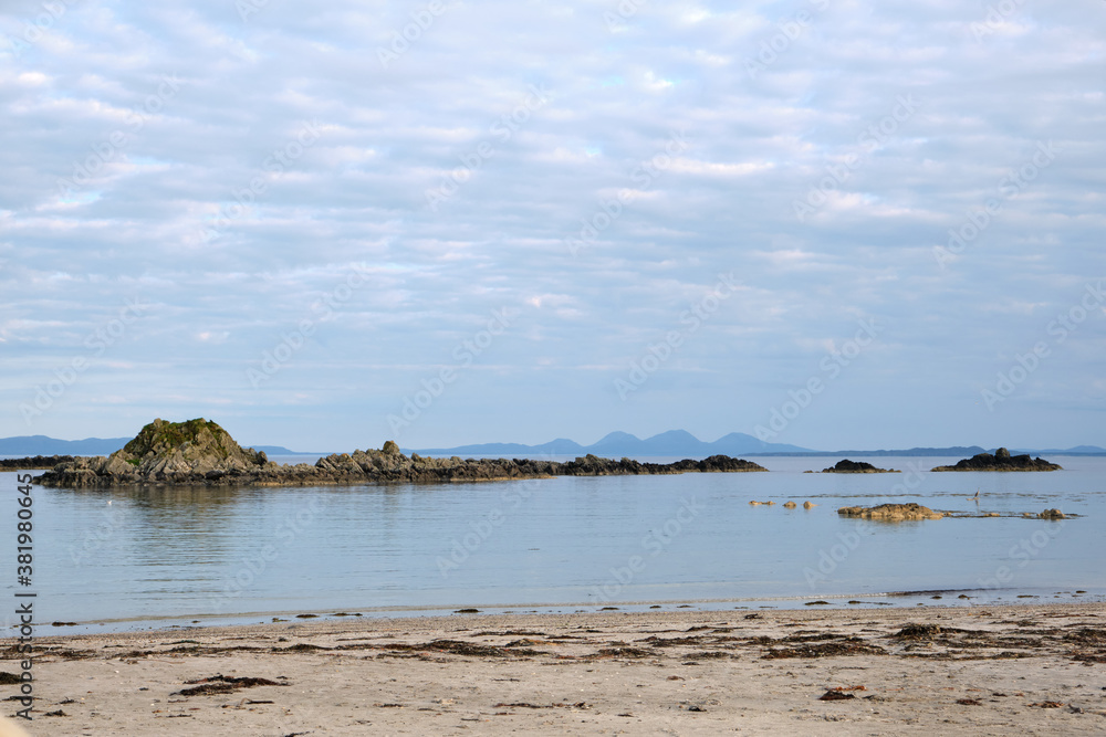 A dramatic view over Uisken Beach out to the Atlantic Ocean with small islands in the foreground and the Isle of Jura mountains visible in the distance. Relaxing view from the Isle of Mull, Scotland. A dramatic view over Uisken Beach out to the Atlantic Ocean with small islands in the foreground and the Isle of Jura mountains visible in the distance. Relaxing view from the Isle of Mull, Scotland.