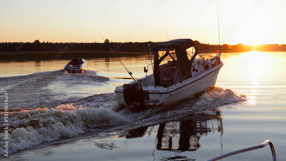 Two outboard motor boats start on beautiful sunset background in sunshine, foam wake tracks in calm water on Sun on Horizon at Sunny summer evening, view from powerboat, outdoor active recreation