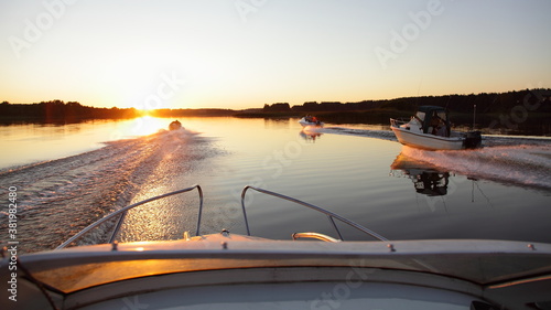 Fast gliding 3 motor boats on beautiful sunset background in sun shine, foam wake tracks in calm water on forest stripe on Horizon at Sunny summer evening, front view from powerboat bow deck railing