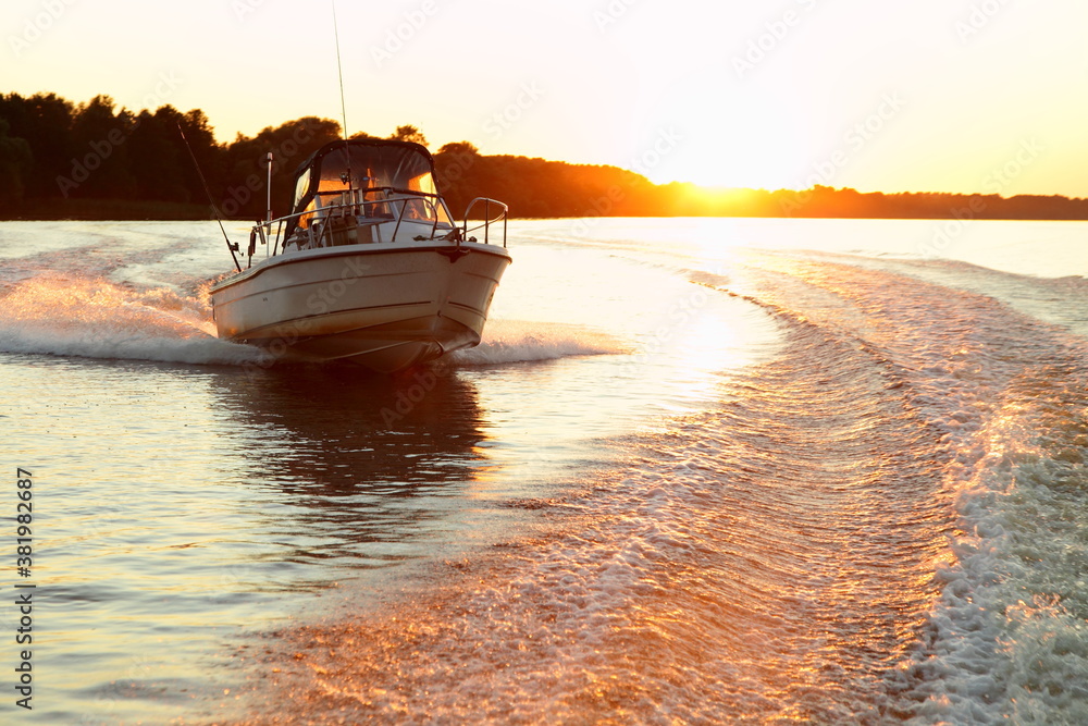 Fast gliding motor boat in wake track on beautiful sunset background, bright orange sun shine in calm water on forest stripe on Horizon at Sunny summer evening, front side view from powerboat stern - obrazy, fototapety, plakaty 