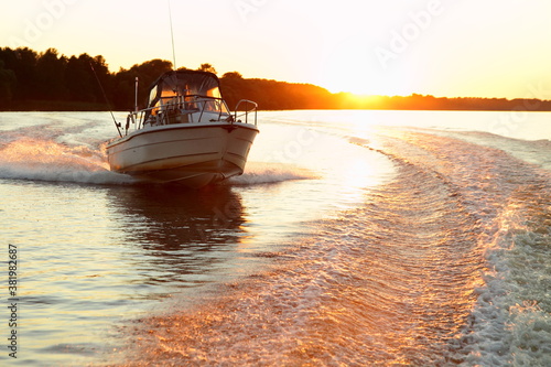 Obraz na płótnie Fast gliding motor boat in wake track on beautiful sunset background, bright orange sun shine in calm water on forest stripe on Horizon at Sunny summer evening, front side view from powerboat stern w salonie
