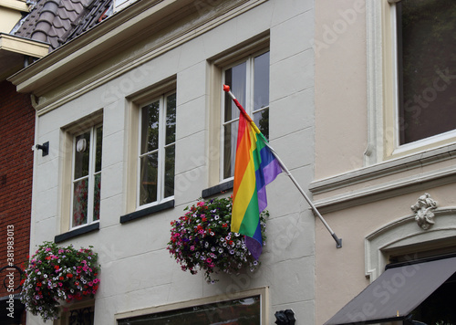 Flagge mit Regenbogenfarben auf einem weißen Haus in den Niederlanden. An der Wand hängen zwei Blumenkörbe.