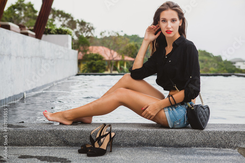 young sexy beautiful woman sitting at pool barefoot, long skinny tanned legs, high heeled shoes, black shirt, denim shorts, purse, relaxing, summer style, vacation, close-up details © mary_markevich