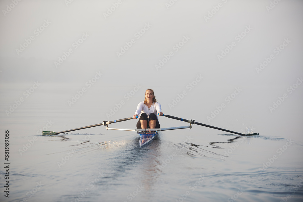 Middle aged woman rowing on lake at early morning with mist over water.