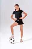 Full-length shot of a cute teenage girl smiling at camera while exercising with soccer ball, isolated over grey background in studio