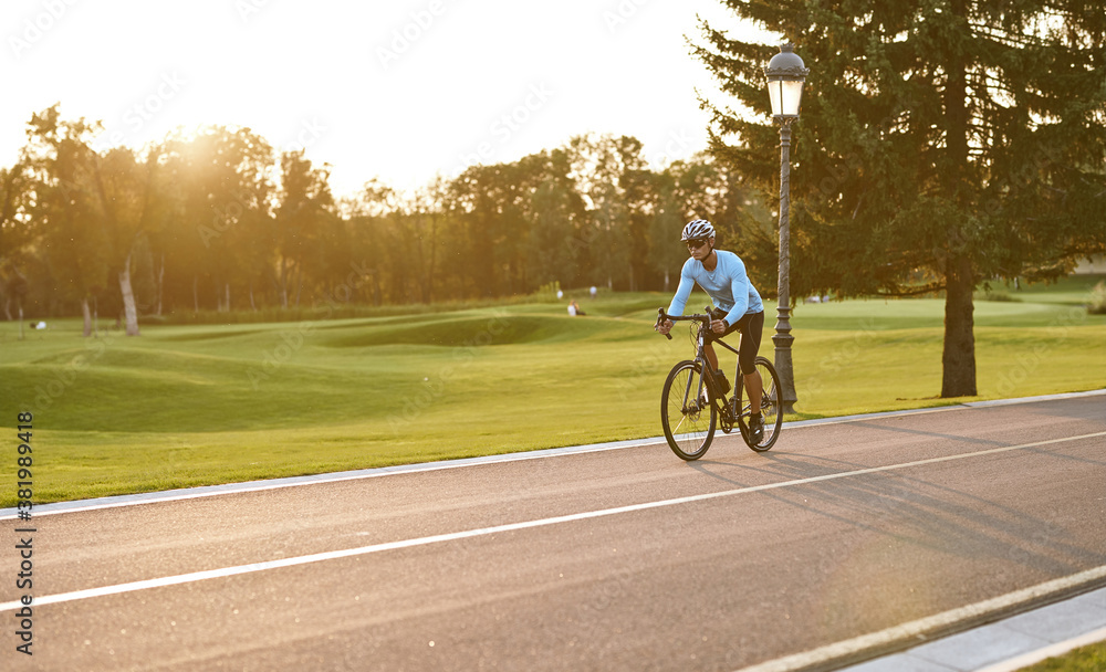 Athletic man in sportswear cycling in city park at sunset, riding bicycle along a road