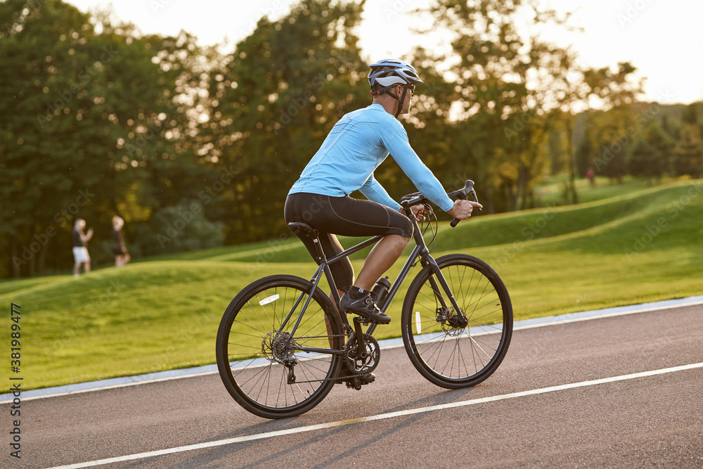 Side view of athletic man in sportswear riding bicycle along a road in city park at sunset Side view of athletic man in sportswear riding bicycle along a road in city park at sunset
