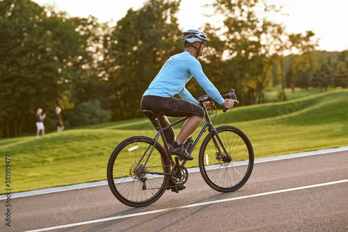 Side view of athletic man in sportswear riding bicycle along a road in city park at sunset