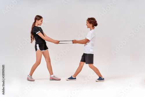 Full-length shot of happy teenage boy and girl having fun while exercising with resistance band, standing isolated over grey background