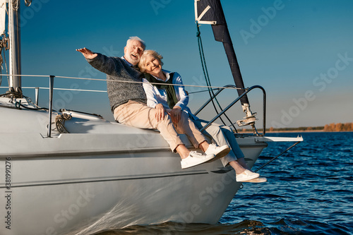 Beautiful happy elderly couple relaxing on sail boat or yacht deck floating in a calm blue sea, they looking at the horizon and enjoying amazing sunset © Kostiantyn