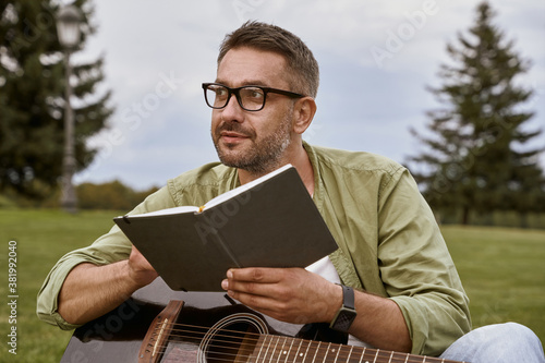 Young thoughtful man wearing eyeglasses holding acoustic guitar and composing a song while sitting on a green grass outdoors, male musician working in park