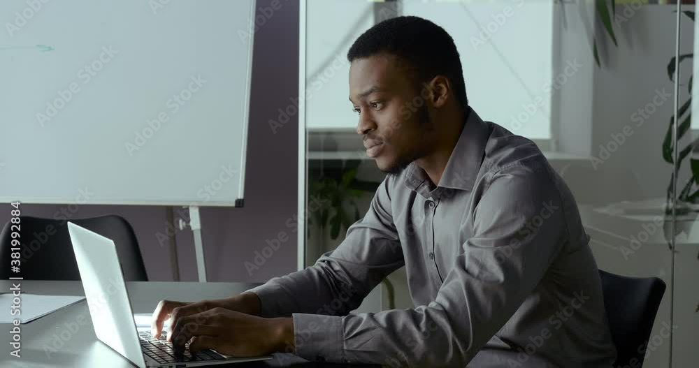 Portrait of one young African man typing on laptop sitting in office writing notes in notebook, serious american businessman working with technology indoor, black adult student study online in net