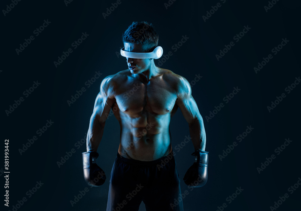 Muscular model sports young man in glasses of virtual reality and boxing gloves on dark background. Fashion portrait of strong brutal guy. Sexy torso. Male flexing his muscles. VR. Blue neon light. Muscular model sports young man in glasses of virtual reality and boxing gloves on dark background. Fashion portrait of strong brutal guy. Sexy torso. Male flexing his muscles. VR. Blue neon light.