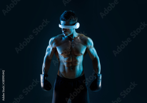 Muscular model sports young man in glasses of virtual reality and boxing gloves on dark background. Fashion portrait of strong brutal guy. Sexy torso. Male flexing his muscles. VR. Blue neon light.