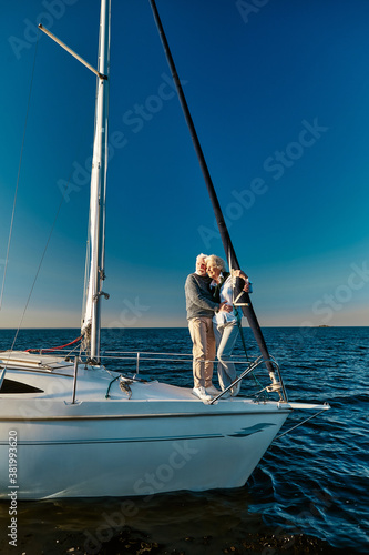 In love with the sea. Full length of beautiful senior couple standing on the side of sail boat or yacht deck floating in the calm blue sea, hugging and enjoying amazing sunset