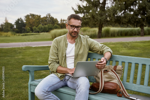 Young handsome man sitting on the wooden bench in park and using laptop, working outdoors © Kostiantyn