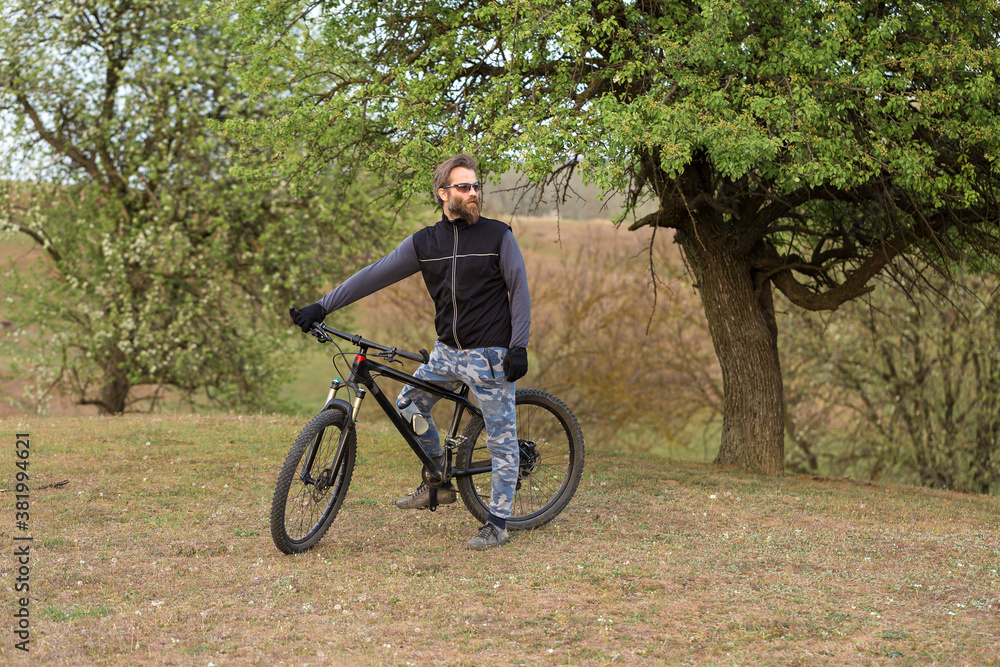Obraz premium Sports brutal bearded guy on a modern mountain bike. Cyclist on the green hills in the spring.
