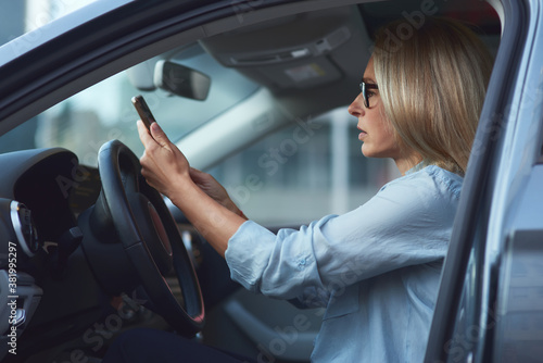 Side view of a concentrated woman or business lady wearing eyeglasses sitting behind steering wheel in her modern car and using smartphone