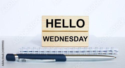 The text Hello Wednesday on a bar of jenga wood, lying on a Notepad with a metal blue pen