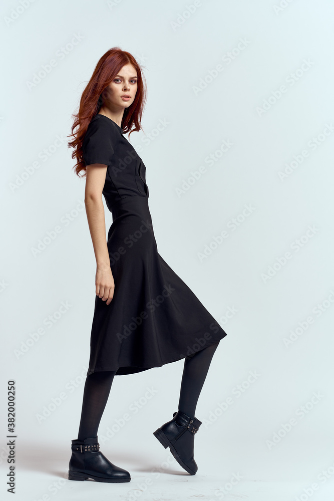 A woman in a black dress on a light background and pantyhose shoes red hair and pose in full growth A woman in a black dress on a light background and pantyhose shoes red hair and pose in full growth