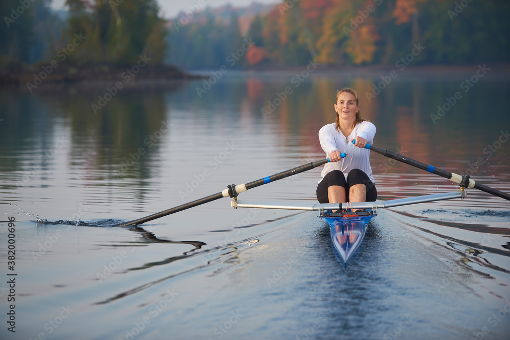 Middle aged woman rowing on a calm lake at sunrise with autumn colours in background. Middle aged woman rowing on a calm lake at sunrise with autumn colours in background.