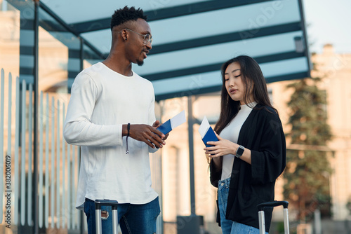 African-american man and asian woman holding passports and talking at bus stop.