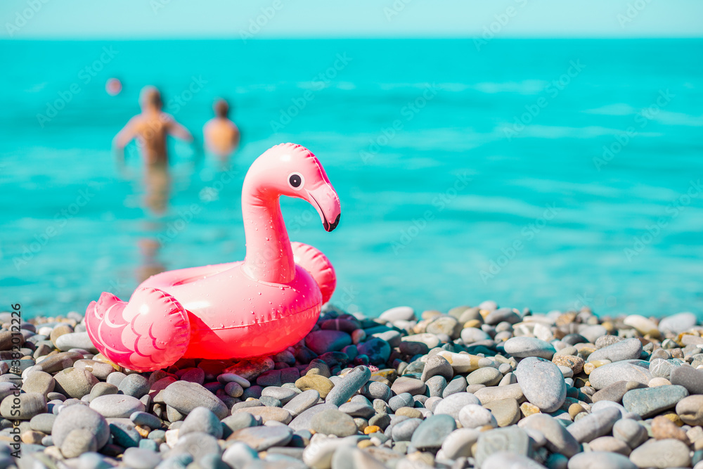 Fototapeta Inflatable toy for playing on the water - pink Flamingo on the sea stone shore, on the background - people swimming in the sea