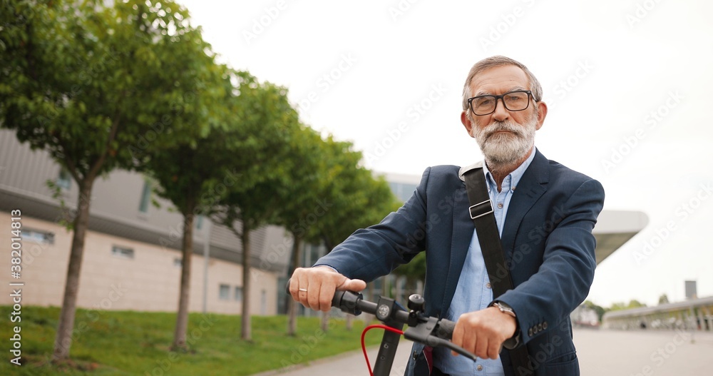 Portrait shot of Caucasian old man with gray hair and in glasses sitting on bike and looking at camera. Senior gray-haired grandfather on electric scooter outdoors. Male pensioner on bicycle in city.