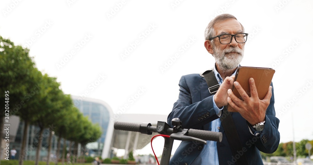 Senior gray-haired man in glasses and headphones standing at bike on street and tapping or scrolling on gadget computer. Old grandfather in eyeglasses using tablet device and browsing online outdoor. Senior gray-haired man in glasses and headphones standing at bike on street and tapping or scrolling on gadget computer. Old grandfather in eyeglasses using tablet device and browsing online outdoor.
