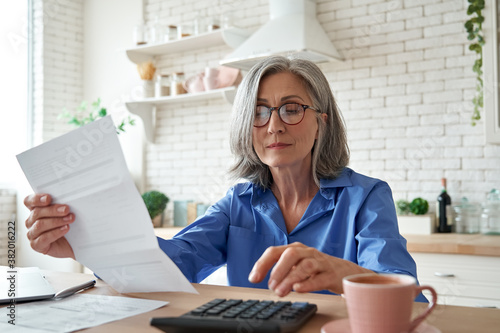 Senior mature business woman holding paper bill using calculator, old lady managing account finance, calculating money budget tax, planning banking loan debt pension payment sit at home kitchen table. photo