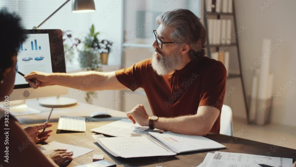 Mature businessman with grey beard pointing at diagram on computer screen and discussing project with afro-american female colleague while working together in office