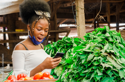 young pretty african businesswoman standing in the market got surprise about what she saw on her phone while wearing face mask to prevent herself from the outbreak in the society.