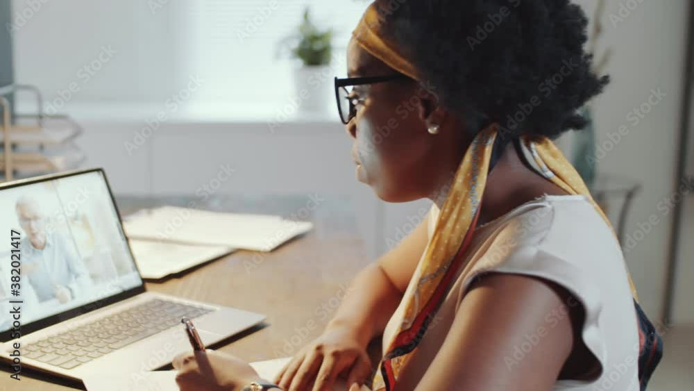 Young African American woman speaking with senior male business partner via online video call on laptop and taking notes on paper while working in office in the evening Young African American woman speaking with senior male business partner via online video call on laptop and taking notes on paper while working in office in the evening
