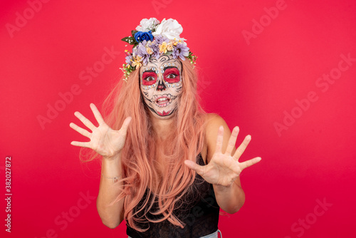young beautiful woman wearing halloween make up over red studio background, afraid and terrified with fear expression stop gesture with hands, shouting in shock. Panic concept.