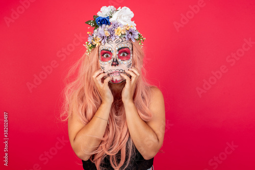 Young beautiful woman wearing halloween make up over red background keeps hands near mouth, feels frightened and scared, has a phobia, Shock and frighted concept.