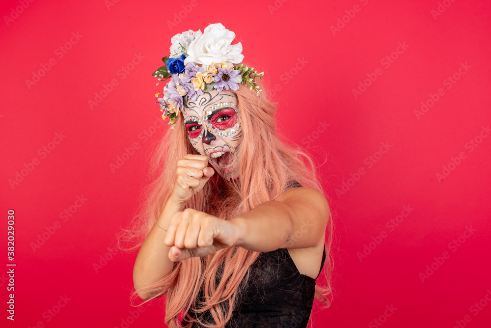 young beautiful woman wearing halloween make up over red punching air with fist and looking confidently at camera, male struggle, fighting spirit. young beautiful woman wearing halloween make up over red punching air with fist and looking confidently at camera, male struggle, fighting spirit.
