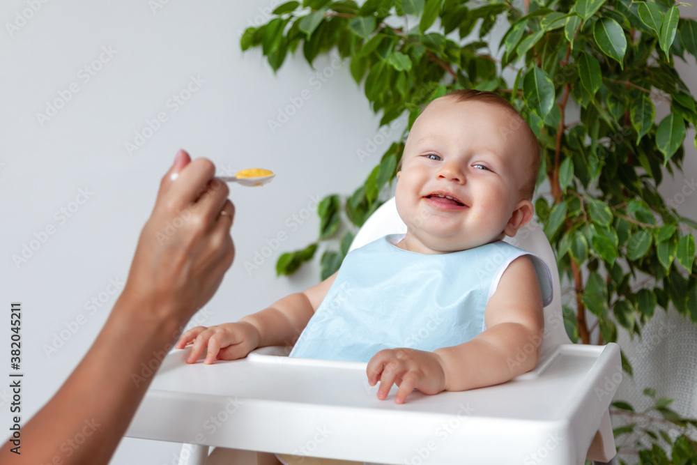 Mother feeds funny happy blond baby in blue bib from a spoon Mother feeds funny happy blond baby in blue bib from a spoon